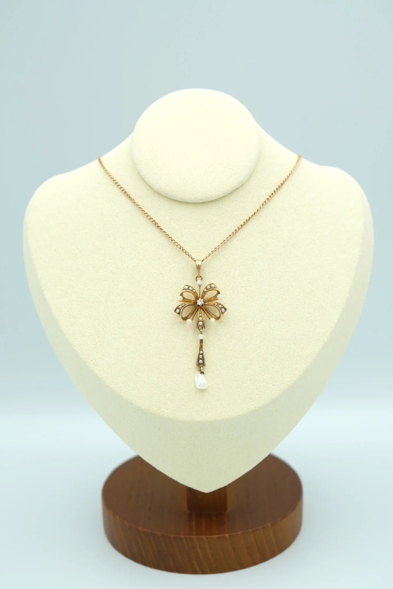 A gold bow pendant hangs on a display bust with a pearl and diamond drop below the pendant, in front of a gray background